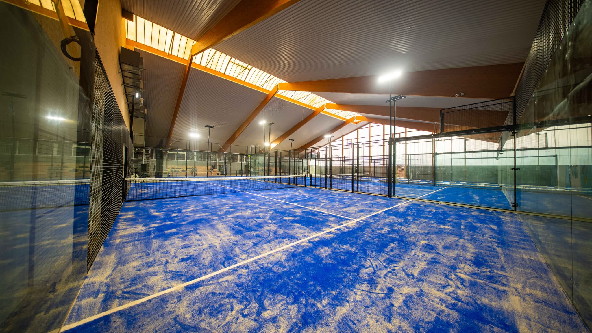Indoor-Padel-Court mit Glaswänden und blauem Belag unter einer Holzdachkonstruktion
