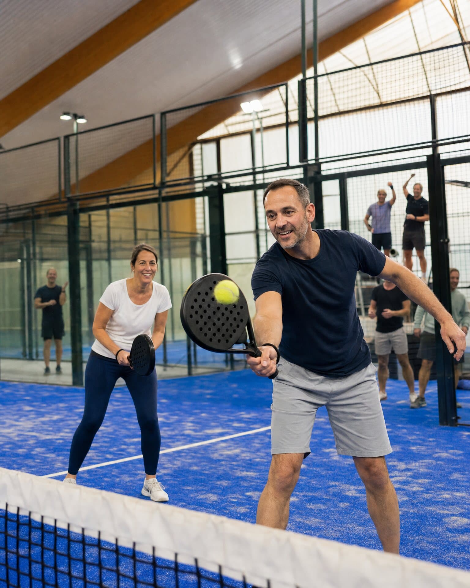Kollegen spielen am Netz ein Doppel mit Padelschlägern, im Hintergrund jubeln weitere Teammitglieder hinter der Glaswand.